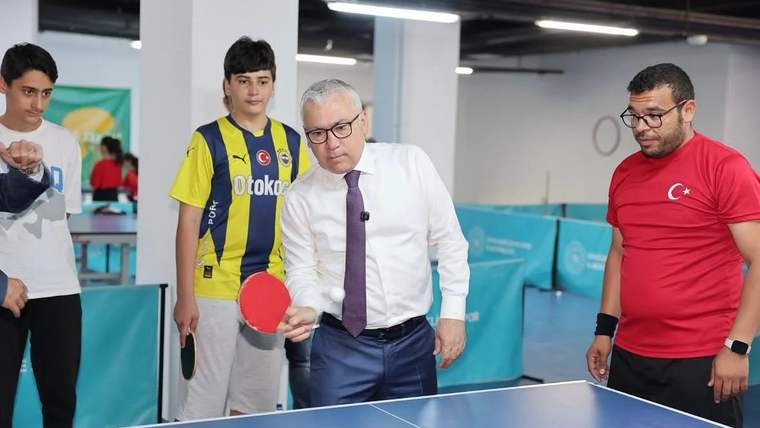 Masa tenisi oynayan bir grup genç ve bir kişi, masanın arkasında raket tutuyor.