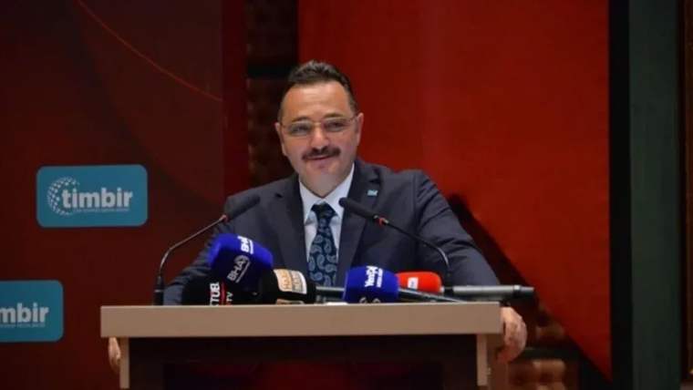 Kürsüde konuşan bir kişi, mikrofonların önünde gülümseyerek mesaj veriyor.