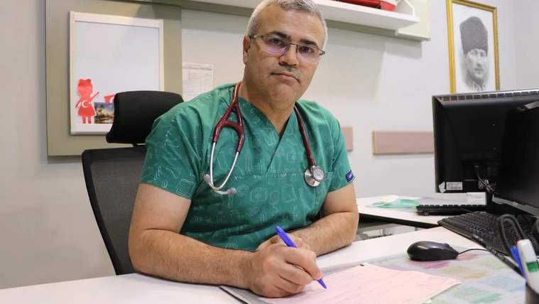 Yeşil önlük giymiş bir doktor, masasında oturarak not alıyor.