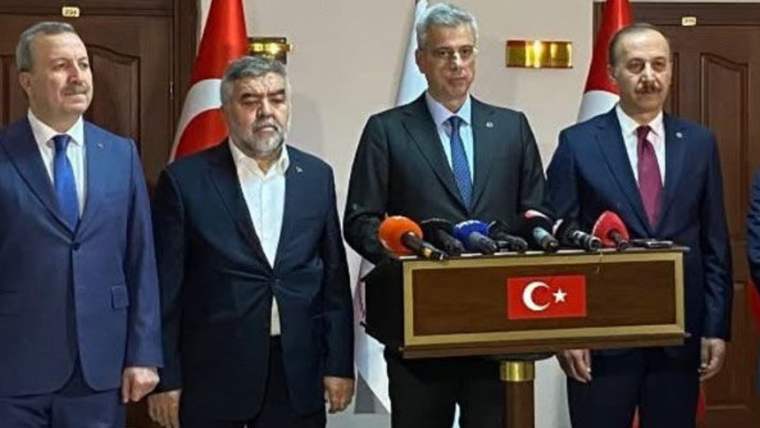 Dört kişi, Türk bayraklarının önünde bir masanın arkasında duruyor. Masada mikrofonlar var.