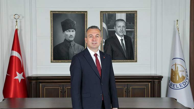 Takım elbiseli bir kişi, Türk bayrağı ve belediye flamaları arasında ahşap makam masasının önünde duruyor.