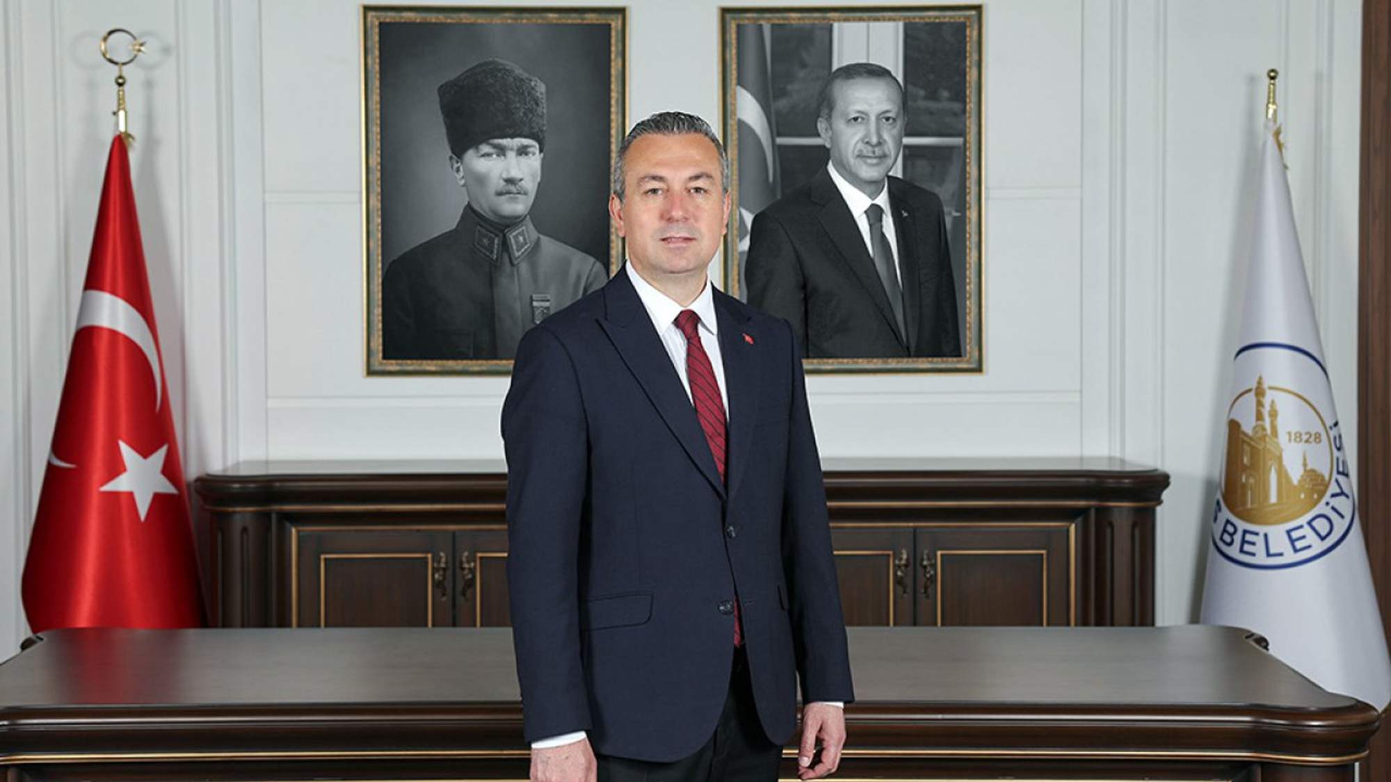 Takım elbiseli bir kişi, Türk bayrağı ve belediye flamalarının bulunduğu odada masanın önünde duruyor.