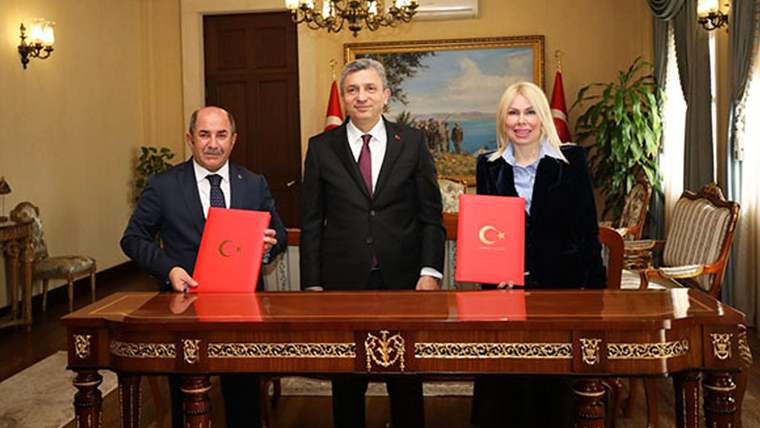 Masada oturan iki kişi kırmızı dosya tutarken, ortada ve arkalarında ayakta duran bir kişi görülüyor.