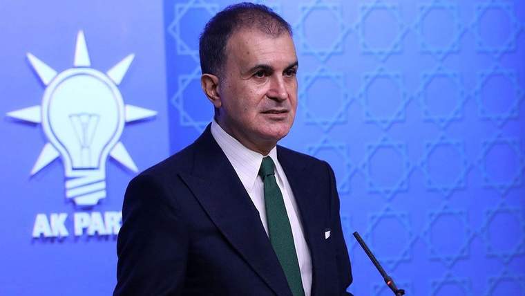 Mikrofonun yanında duran takım elbiseli bir kişi, AK Parti logosu bulunan mavi bir arka plan önünde görülüyor.