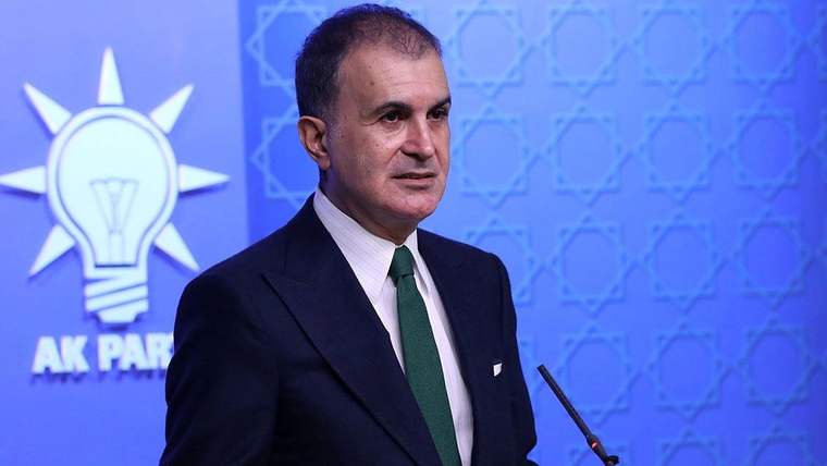 Koyu takım elbiseli bir kişi, mavi fon önünde mikrofon karşısında konuşurken görülüyor.
