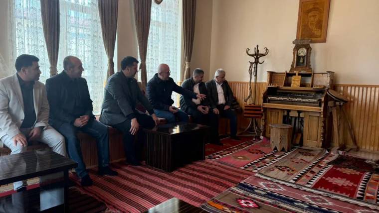 Halılarla döşeli odada, pencere önünde oturan altı erkek ve sağda eski bir piyano görülüyor.