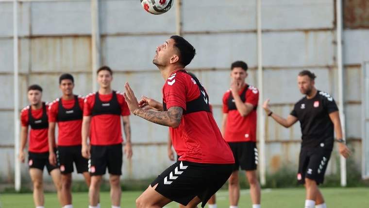 Antrenman sahasında kırmızı formalı bir futbolcu havadaki topa bakarken, diğer futbolcular arka planda duruyor.