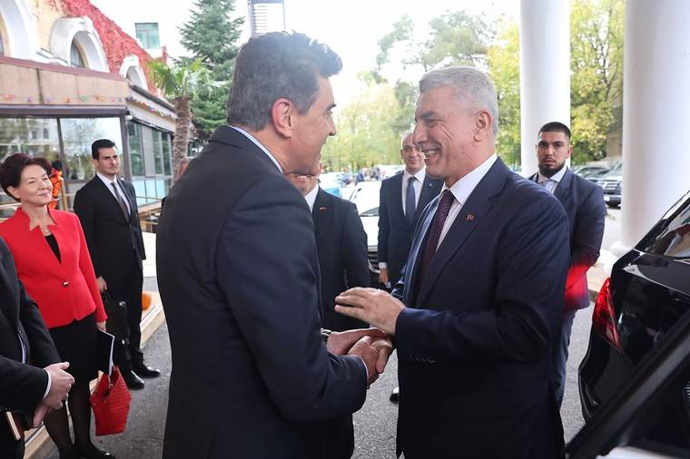 Takım elbiseli iki erkek, siyah bir aracın yanında el sıkışırken çevrelerinde birkaç kişi bekliyor.