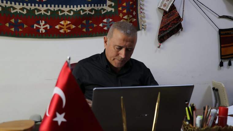 Bir kişi, Türk bayrağı bulunan masada dizüstü bilgisayar kullanırken duvardaki dokuma süsler görülüyor.