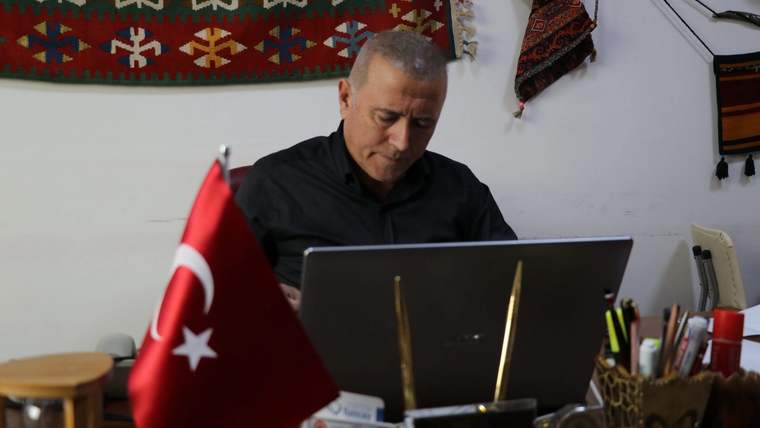 Bir kişi, Türk bayrağı bulunan masada dizüstü bilgisayar kullanırken duvardaki dokuma süsler görülüyor.