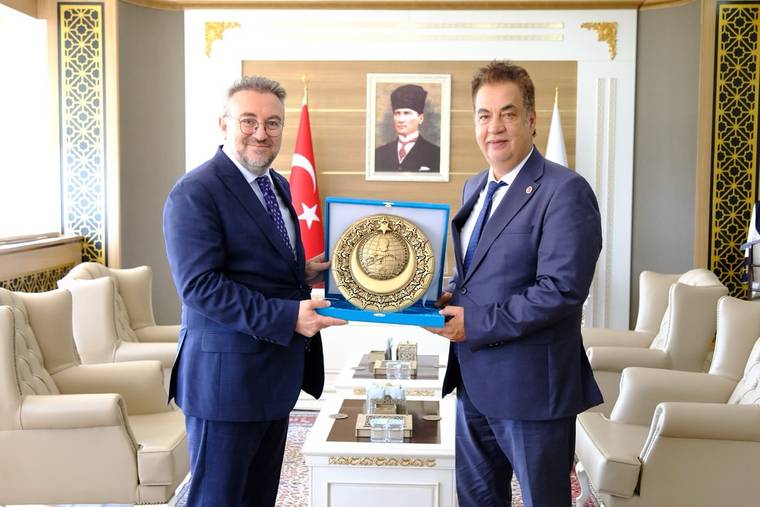Bir odada iki takım elbiseli kişi, üzerinde madalya bulunan mavi plaketi birlikte tutuyor; arkada Türk bayrağı var.