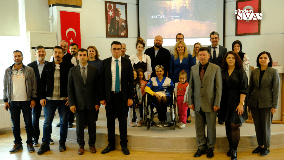 Bir grup insan, sahnede bir araya gelerek fotoğraf çektiriyor. Arka planda Türk bayrağı ve bir ekran var.