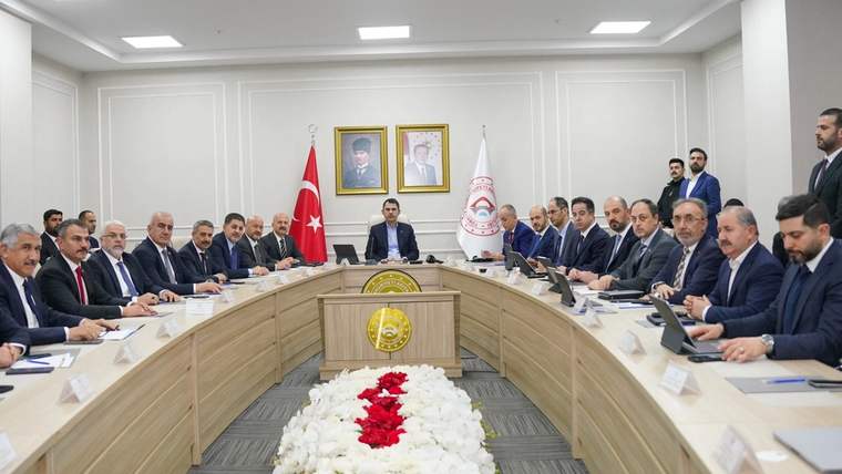 Toplantı masası çevresinde oturan çok sayıda kişi, Türk bayrağı ve çiçeklerle süslü salon görülüyor.