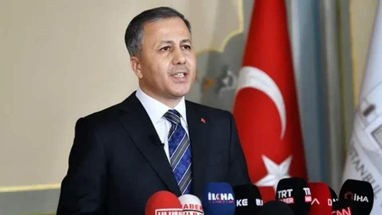 Mikrofonların önünde konuşan takım elbiseli bir kişi, arka planda Türk bayrağıyla görülüyor.