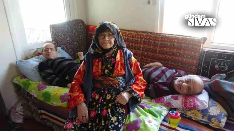 Renkli başörtüsü ve çiçekli kıyafet giyen yaşlı kadın, iki kişinin uzandığı odada oturuyor.