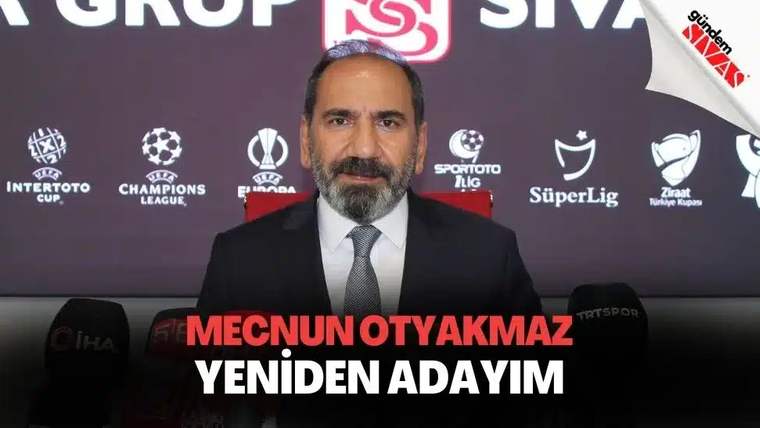 Kırmızı arka planda oturan bir adam, önünde mikrofonlar var ve gülümseyerek kameraya bakıyor.