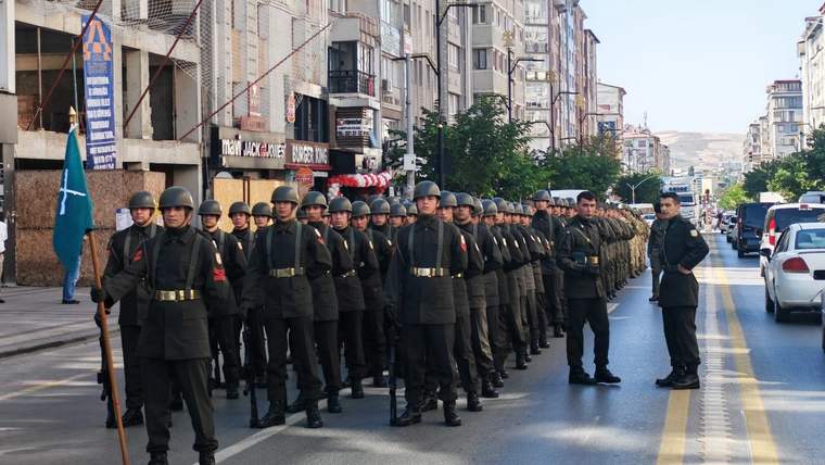 Şehir caddesinde sıralar halinde yürüyen üniformalı askerler, binalar ve araçlar arasında ilerliyor.