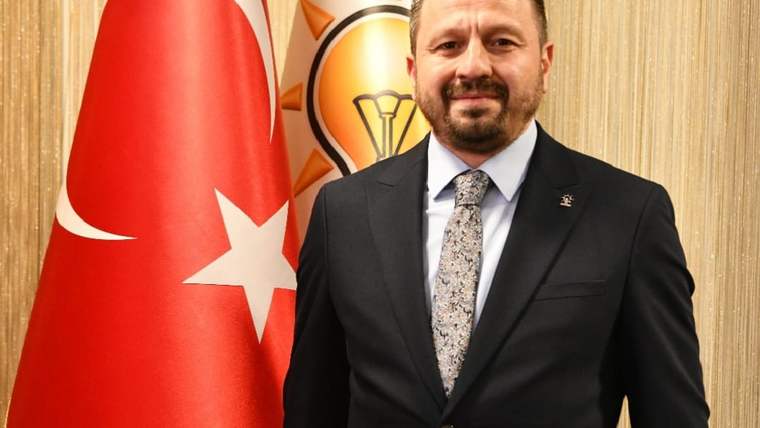 Takım elbiseli ve kravatlı bir kişi, Türk bayrağı ile üzerinde amblem bulunan bir fonun önünde duruyor.