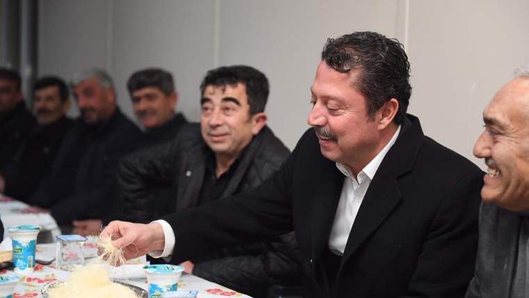 Masada oturan bir grup erkekten biri, tabaktaki yiyecekten eliyle alırken diğerleri onu izliyor.