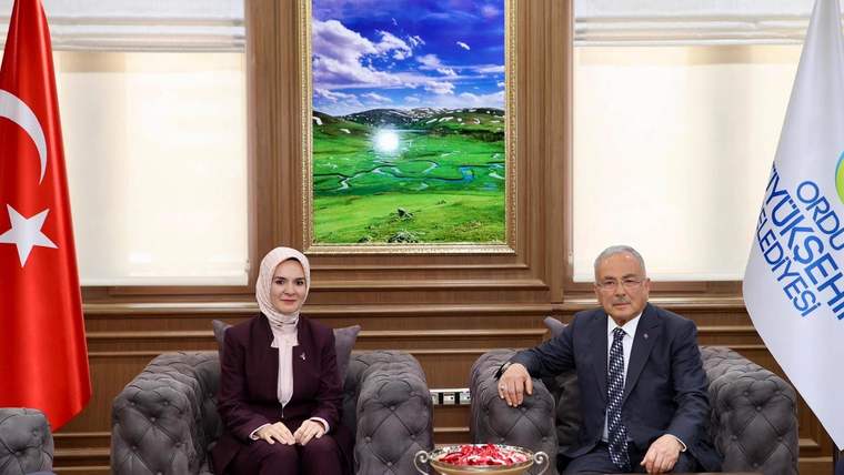 Türk bayrağı ve beyaz bir bayrağın bulunduğu odada, iki kişi koltuklarda oturuyor; önlerinde sehpa var.