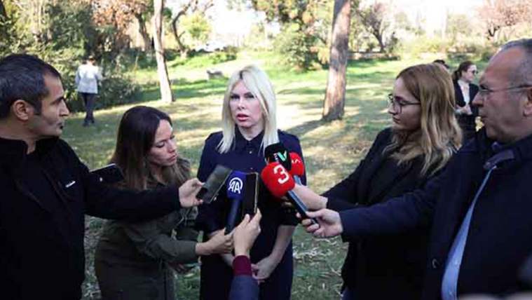 Bir grup gazeteci, bir kadına mikrofon uzatıyor; arka planda ağaçlar ve insanlar görünüyor.