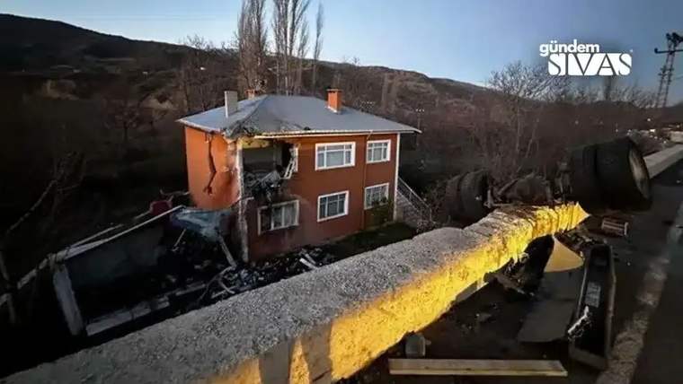 İki katlı turuncu evin üst katında ağır hasar, önünde devrilmiş kamyon ve kırılmış beton bariyer görülüyor.