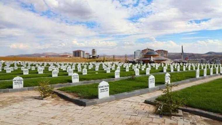 Beyaz mezar taşlarıyla dolu bir mezarlık, arka planda binalar ve bir yapı görünmekte.