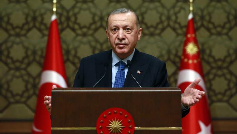 Kürsüde konuşan bir kişi, iki Türk bayrağının önünde mikrofonlarla duruyor.