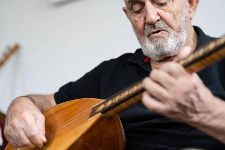 Beyaz sakallı bir kişi, oturarak bağlama çalıyor; elleri sazın gövdesi ve sapı üzerinde görülüyor.