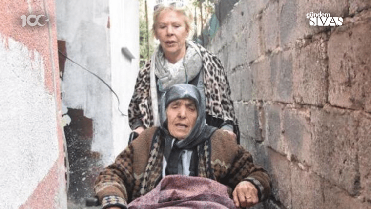 Dar bir geçitte tekerlekli sandalyede oturan yaşlı kadın, arkasında duran başka bir kadınla birlikte görülüyor.