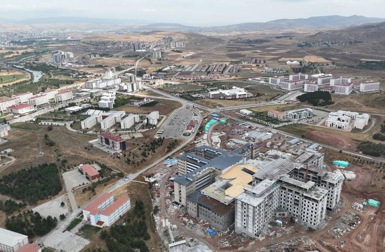 Havadan görünümde, geniş bir yerleşke içinde inşaatı süren büyük ve çok katlı bir bina öne çıkıyor.