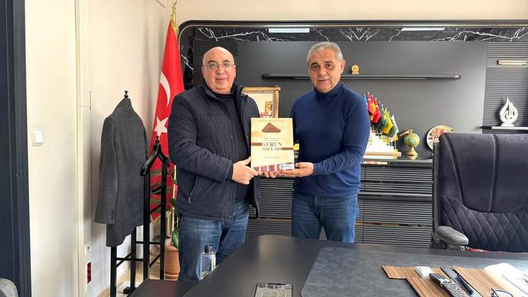İki kişi, bir belgeyi birbirine verirken gülümsüyor; arka planda Türk bayrağı ve çeşitli bayraklar var.