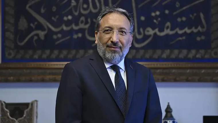 Sakallı bir erkek koyu renk takım elbiseyle duruyor; arkasında Arapça hat yazıları ve dekoratif eşyalar bulunuyor.
