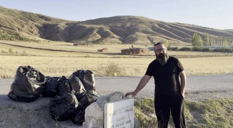 Kırsal alanda siyah çöp torbaları ve beton bir işaretin yanında duran sakallı bir kişi görülüyor.