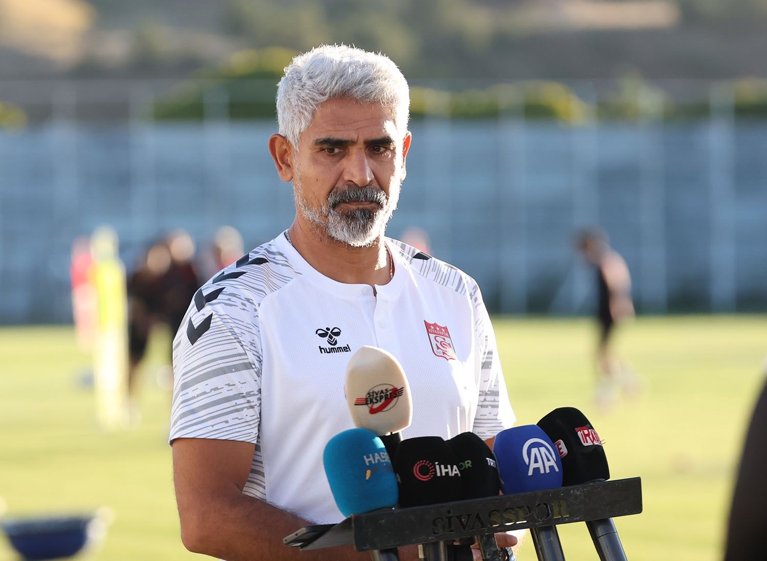 Gri saçlı ve sakallı bir kişi, spor sahasında mikrofonlar önünde konuşurken görülüyor.