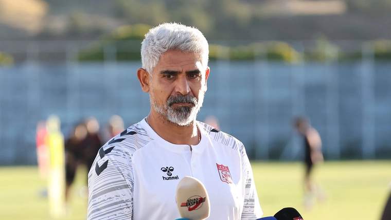Gri saçlı ve sakallı bir kişi, spor sahasında mikrofonlar önünde konuşurken görülüyor.