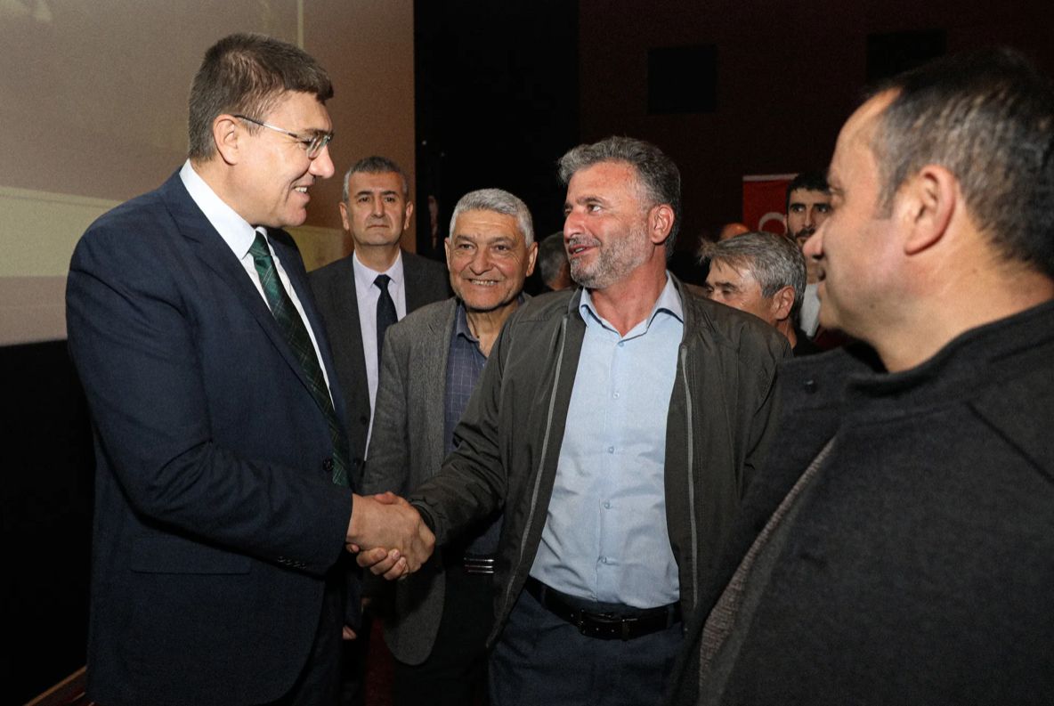Bir grup erkek, bir salonda el sıkışarak sohbet ediyor. Arkada başka insanlar ve bir sahne görünmekte.