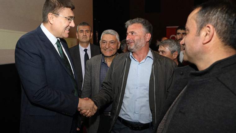 Bir grup erkek, bir salonda el sıkışarak sohbet ediyor. Arkada başka insanlar ve bir sahne görünmekte.