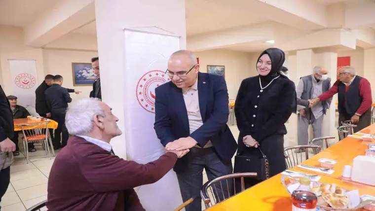Kapalı salonda bir adam, oturan yaşlı bir erkeğin elini sıkarken yanlarında iki kişi duruyor.