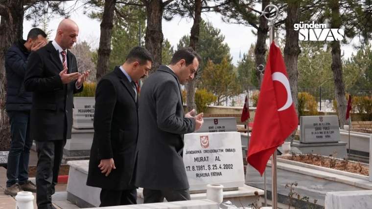 Bir grup erkek, mezar başında dua ediyor; arka planda Türk bayrağı ve ağaçlar var.