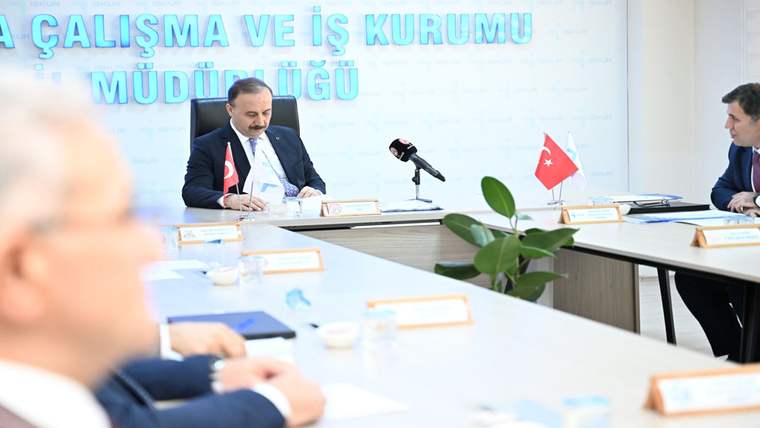 Toplantı masasında oturan bir kişi, mikrofon ve Türk bayraklarının bulunduğu salonda evraklara bakıyor.