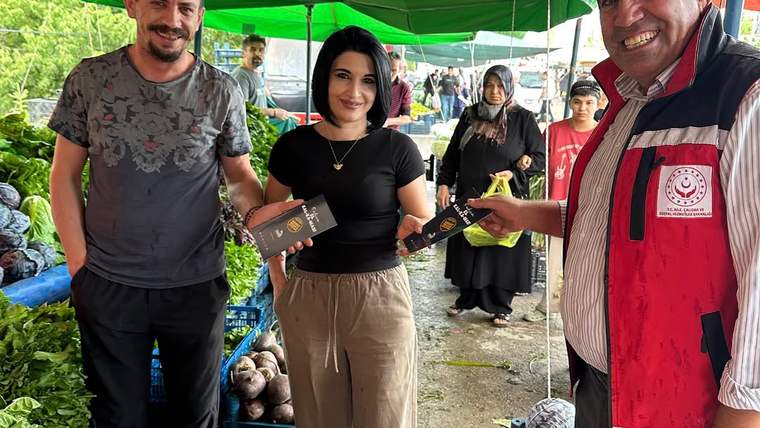 Pazar tezgâhının önünde üç kişi, ellerindeki bilgilendirme broşürlerini kameraya doğru tutuyor.