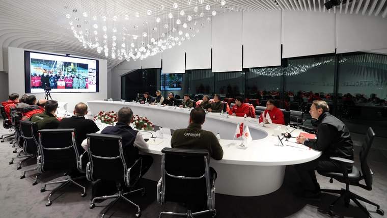 Geniş bir toplantı salonunda, çok sayıda kişi yuvarlak masa çevresinde oturuyor; ekranda sunum görüntüsü var.