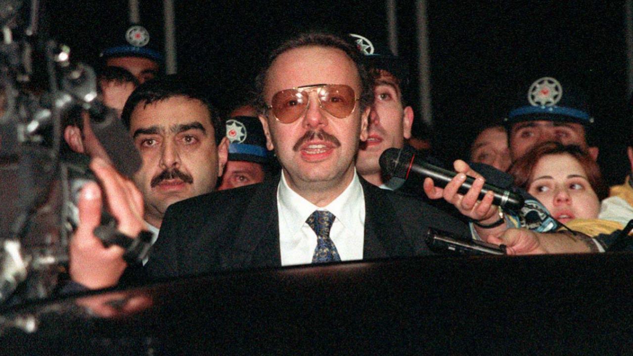 Takım elbiseli bıyıklı bir kişi, mikrofon ve kameralarla çevrili halde kalabalığa konuşuyor.