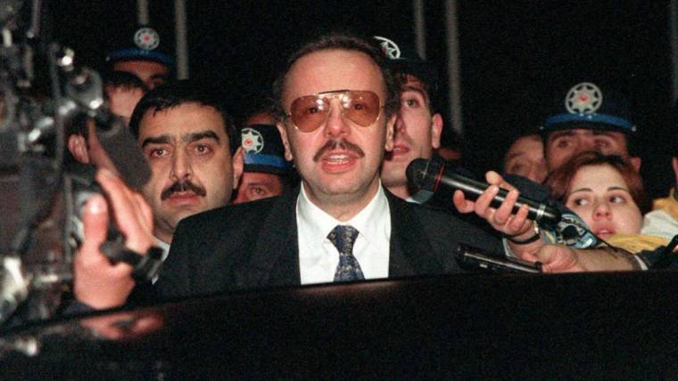 Takım elbiseli bıyıklı bir kişi, mikrofon ve kameralarla çevrili halde kalabalığa konuşuyor.