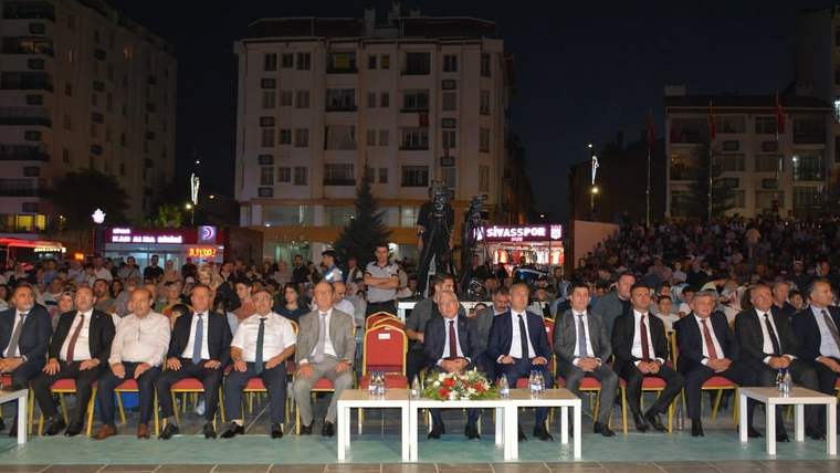 Gece açık alanda, ön sırada oturan takım elbiseli kişiler ve arkalarında kalabalık görülüyor.