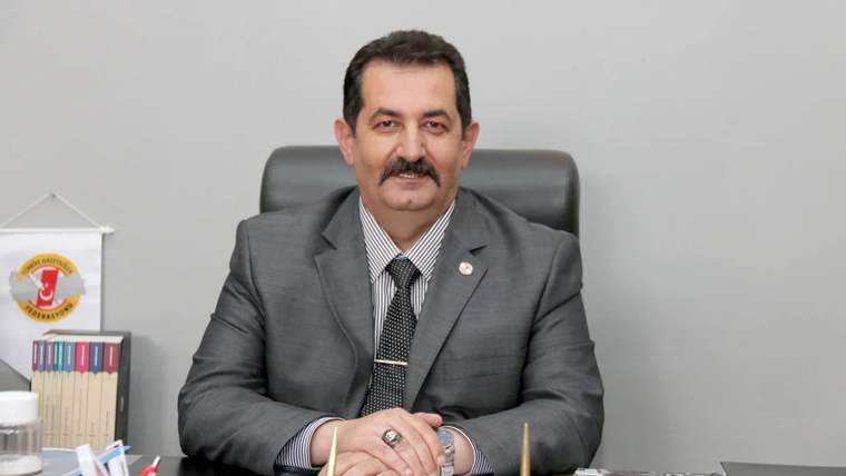 Bir masa başında oturan, takım elbise giymiş bir kişi, arkasında Türk bayrağı ve kitaplar var.
