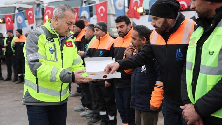 Reflektörlü kıyafetler giyen bir grup görevli, öndeki kişiye beyaz bir kutu uzatırken görülüyor.