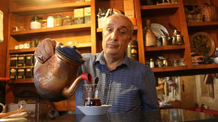 Bir kişi, çaydanlıkla çay dökerken, arka planda ahşap raflarda çeşitli mutfak eşyaları yer alıyor.