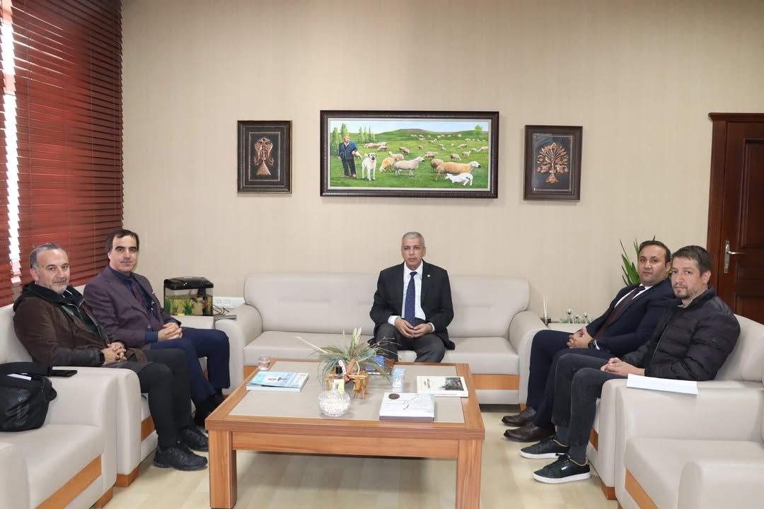Bir odada beş kişi koltuklarda oturuyor; ortadaki kişinin önünde sehpa ve duvarında koyun tablosu bulunuyor.